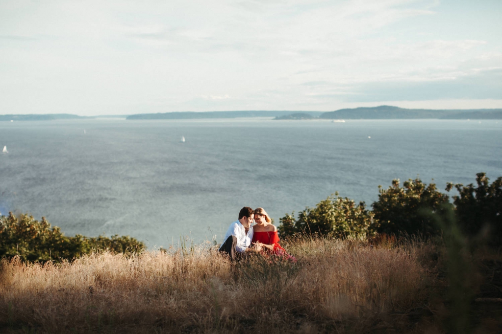engaged couple sitting in a field of tall grass at discovery park in seattle with mountains in the background