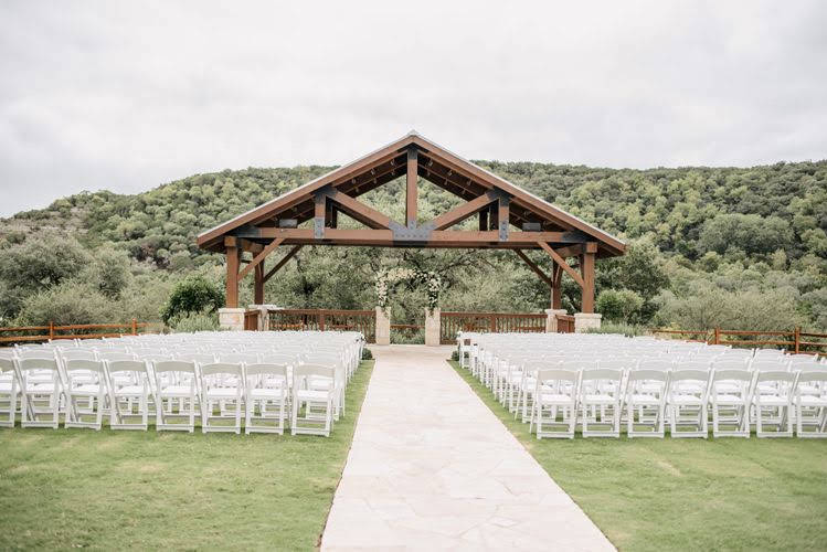 large wooden open-air pavilion at the end of a stone path surrounded by white wedding chairs on a grass lawn
